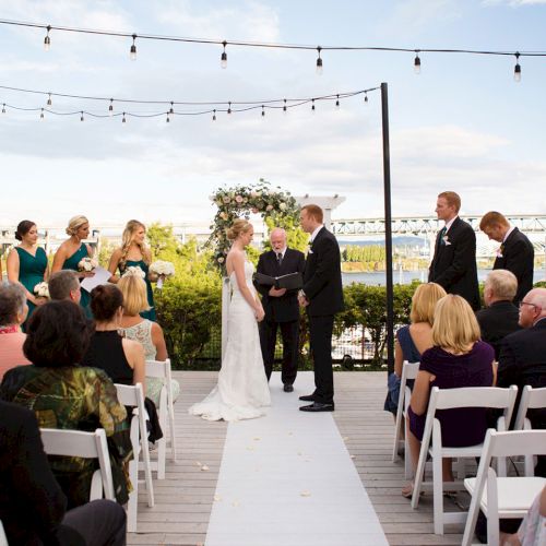 An outdoor wedding ceremony on a wooden deck, with a bride and groom exchanging vows as guests watch beneath string lights.