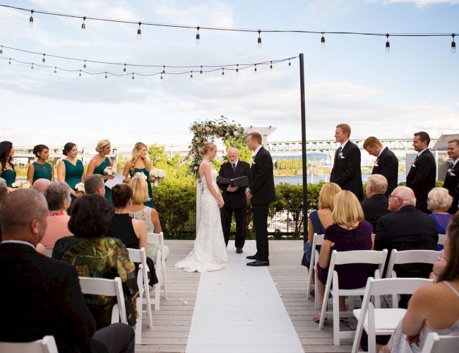 An outdoor wedding ceremony on a wooden deck, with a bride and groom exchanging vows as guests watch beneath string lights.