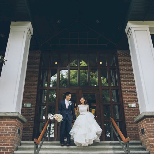 A bride and groom stand on the entrance steps of a brick building with tall white columns and leafy plants, smiling together.