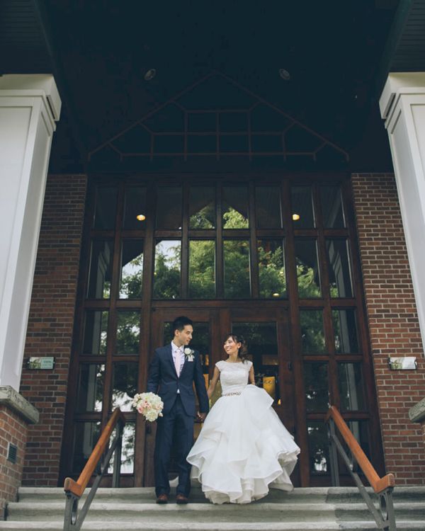 A bride and groom stand on the entrance steps of a brick building with tall white columns and leafy plants, smiling together.