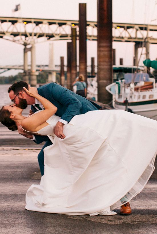 A couple in wedding attire embrace and kiss on a dock by boats, with an industrial waterfront backdrop.