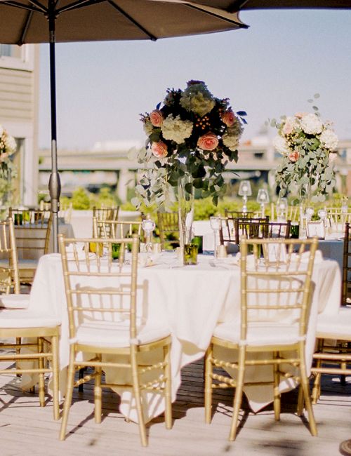 An outdoor event setup with white tablecloths, gold chairs, floral centerpieces, and big patio umbrellas in bright sunshine.