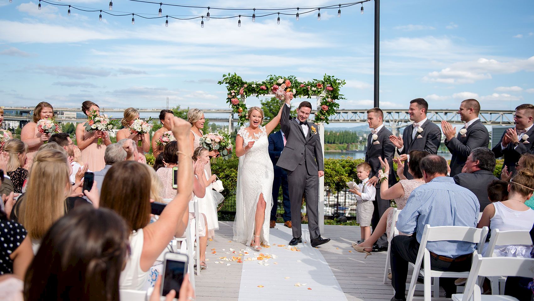 A bride and groom cheer under a floral arch as friends and family clap at an outdoor wedding ceremony by a river.