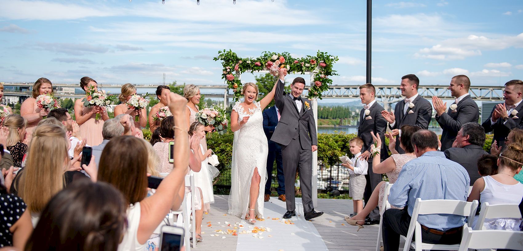 A bride and groom cheer under a floral arch as friends and family clap at an outdoor wedding ceremony by a river.