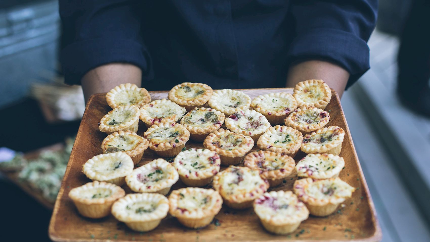 A chef holds a wooden tray of mini quiches with herbs and purple bits, fresh-baked and ready to eat.