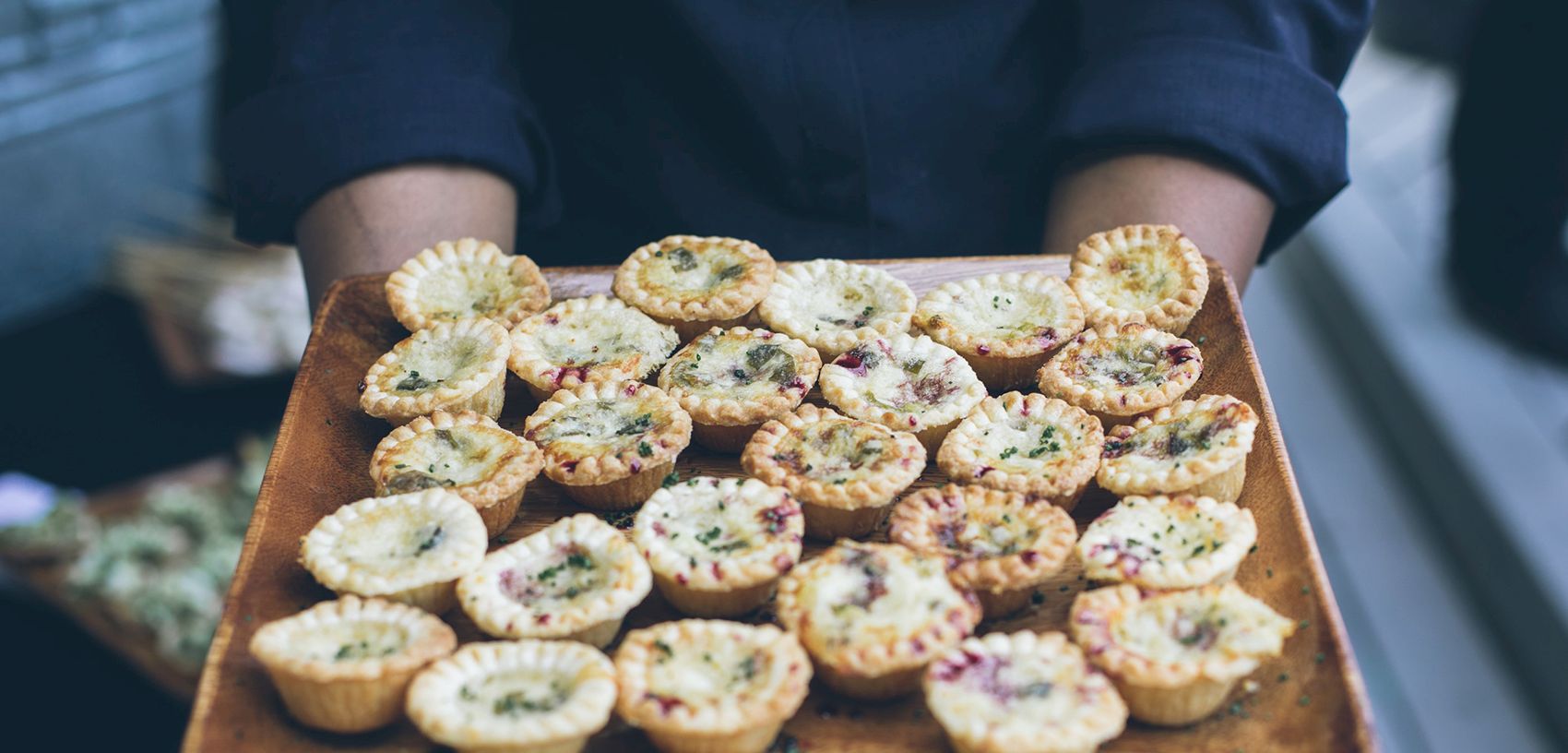 A chef holds a wooden tray of mini quiches with herbs and purple bits, fresh-baked and ready to eat.