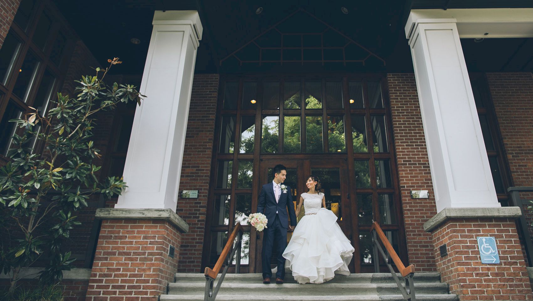 A bride and groom pose on the steps of a brick building, framed by tall columns, flowers, and large doors.