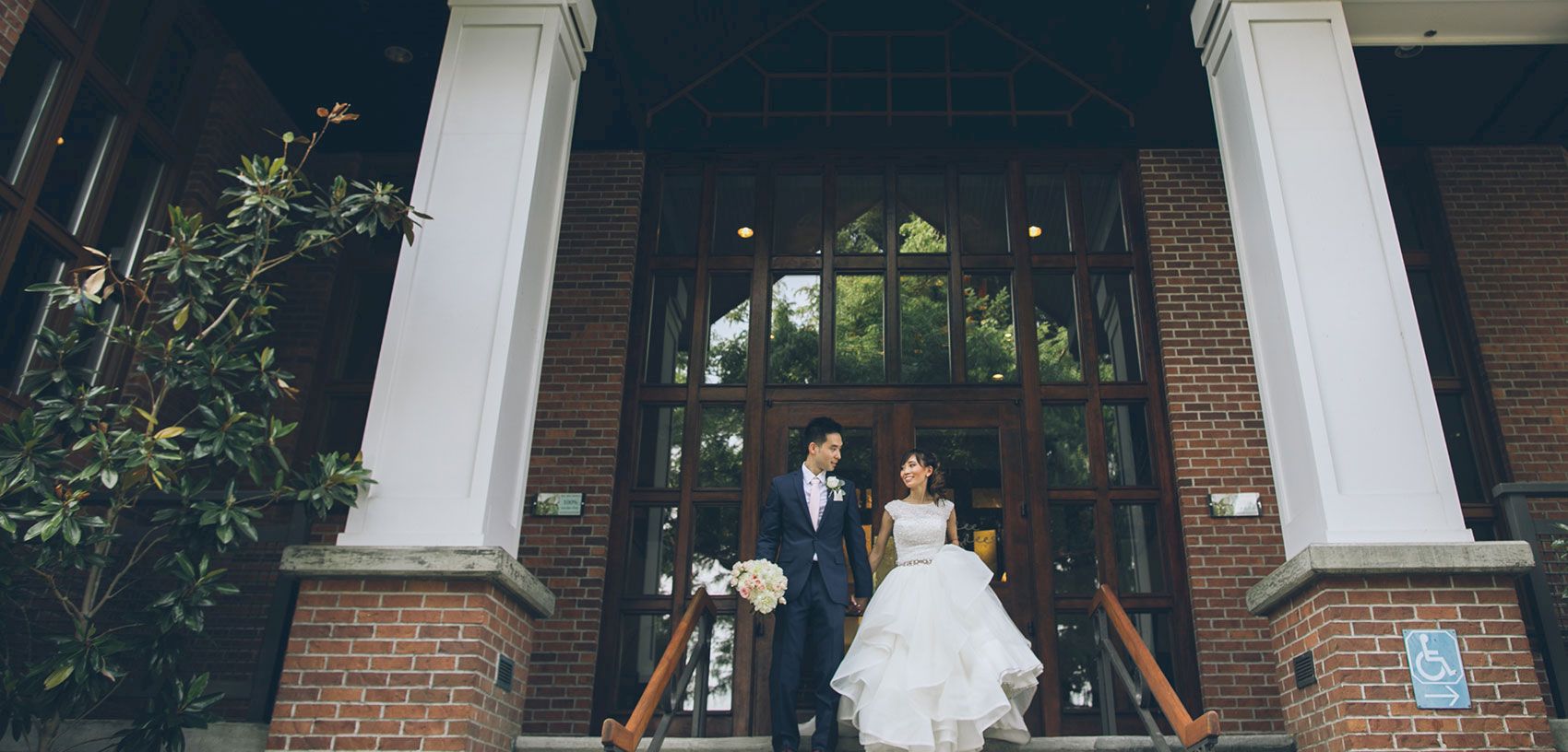 A bride and groom pose on the steps of a brick building, framed by tall columns, flowers, and large doors.