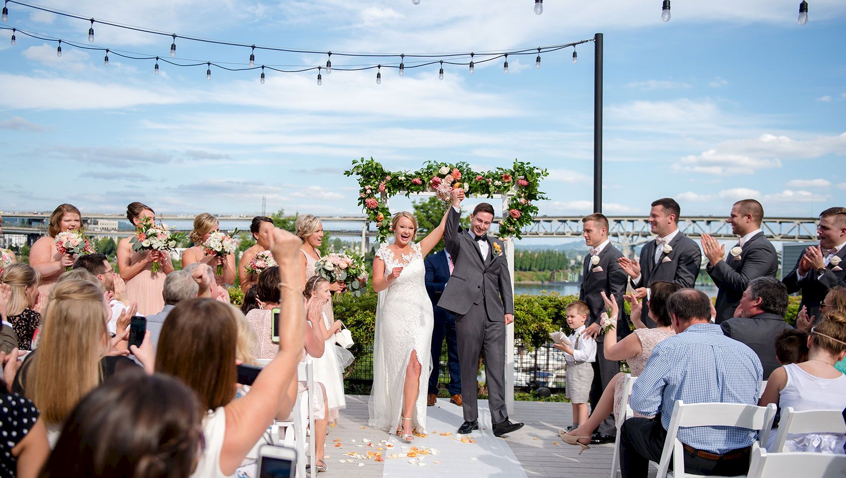 A bride and groom celebrate under a floral arch while guests cheer at an outdoor waterfront wedding ceremony, under string lights.