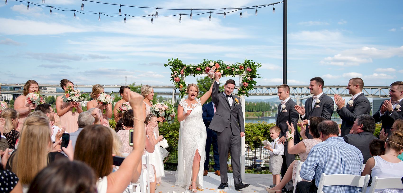 A bride and groom celebrate under a floral arch while guests cheer at an outdoor waterfront wedding ceremony, under string lights.