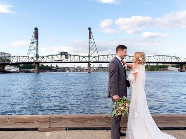 A bride and groom stand by a waterfront railing beside a river and a steel bridge, smiling as they pose for photos.