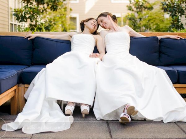 A bride and groom in white wedding dresses lounge on a blue outdoor couch, cuddling in the sunshine, outdoors.