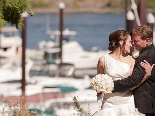 A wedding couple in formal attire share a hug outdoors by a waterfront, with the bride holding a bouquet near an altar.