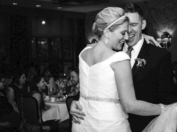 A bride in a white dress and veil dances with a groom in a dark suit at a reception, smiling at each other.