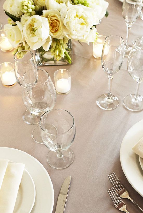 An elegant wedding-style table set with white plates, folded napkins, many wine glasses, and a white flower centerpiece.