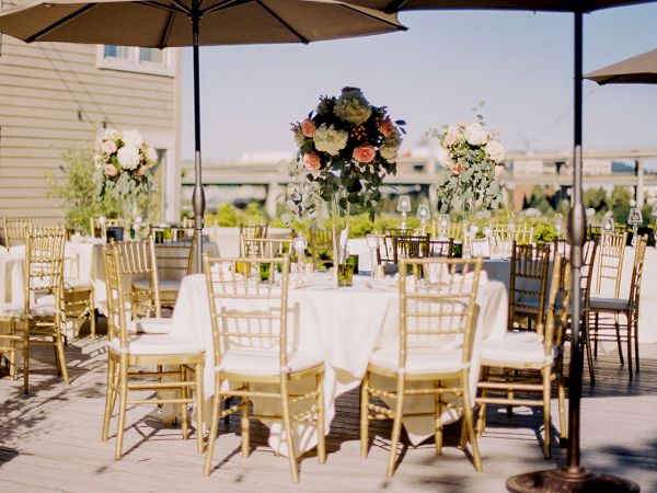 An outdoor wedding reception setup with white tablecloths, gold chairs, and a floral centerpiece under umbrellas.