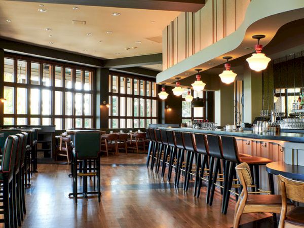 A stylish bar interior with high stools, pendant lights, large windows, and neatly lined bottles behind the counter.