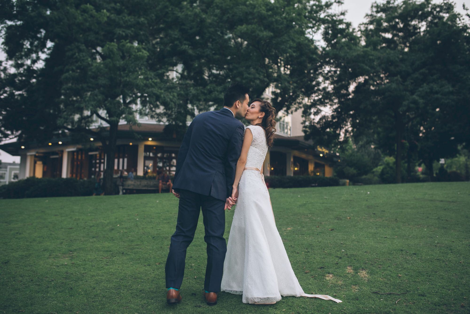 A bride and groom in wedding attire kiss while holding hands on a grassy lawn before a garden building.