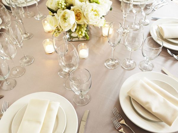 A dressed banquet table with white plates, folded napkins, many wine glasses, candles, and a white flower centerpiece.