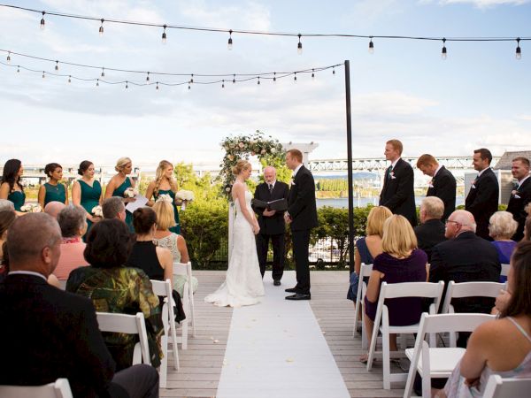 An outdoor wedding ceremony on a terrace, with the bride and groom at the aisle under string lights, surrounded by seated guests.