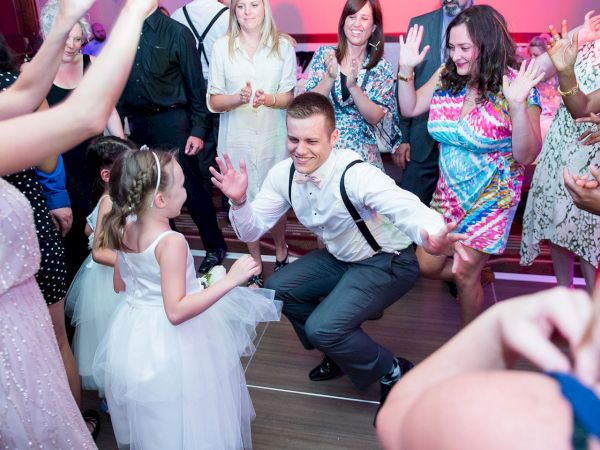 A celebratory dance scene at a party or wedding, with a man kneeling and a little girl holding hands as others cheer and clap.