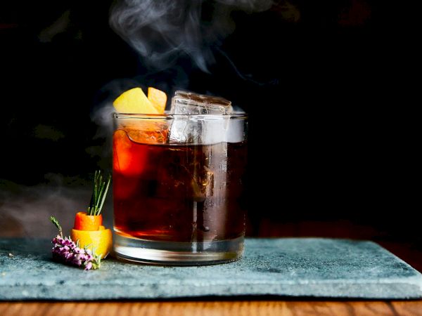 A steaming glass mug of amber tea with apple/orange slices, garnished with herbs on a dark table.