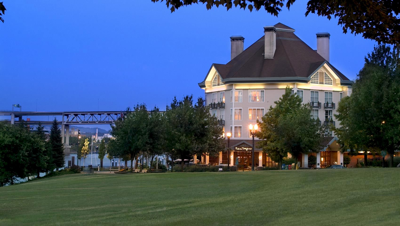 A riverside hotel at dusk with bright lamps, surrounding trees, and a manicured lawn under a clear blue sky.