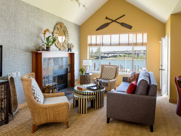 A cozy living room with a fireplace, woven chairs, striped coffee tables, and a large window overlooking a river and bridge.