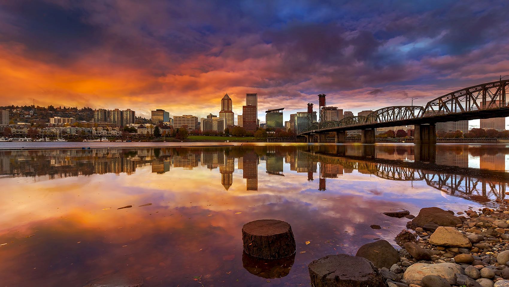 A colorful sunset over a city skyline beside a calm river, with a steel bridge stretching across the water, and rocky shoreline in front.
