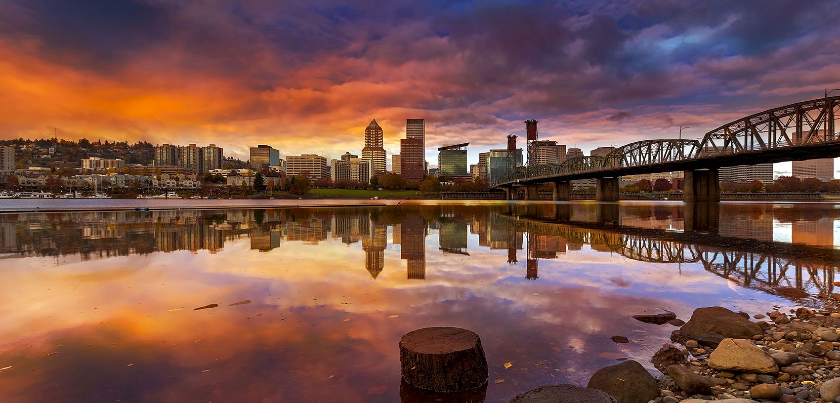 A colorful sunset over a city skyline beside a calm river, with a steel bridge stretching across the water, and rocky shoreline in front.