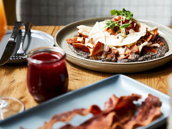 A bowl of open-faced fries or potato hash topped with creamy sauce, mushrooms, tomatoes, and herbs, beside ketchup and utensils.