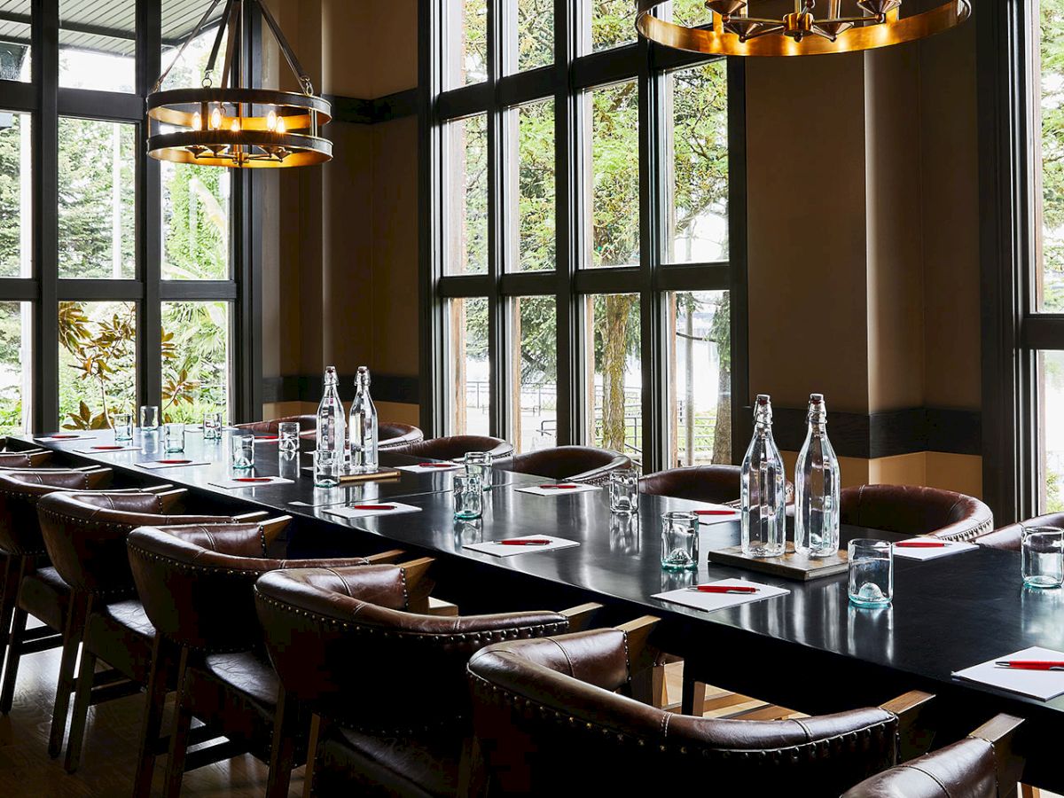 A modern restaurant or event room with a long metal-topped table, leather chairs, bottles, and warm pendant lights by large windows.