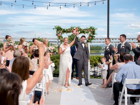 A wedding ceremony outdoors as a bride and groom stand under a floral arch while guests cheer and toss flower petals ending.