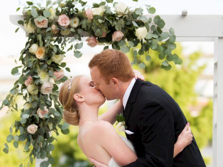 A couple kiss under a lush floral arch of eucalyptus and roses.