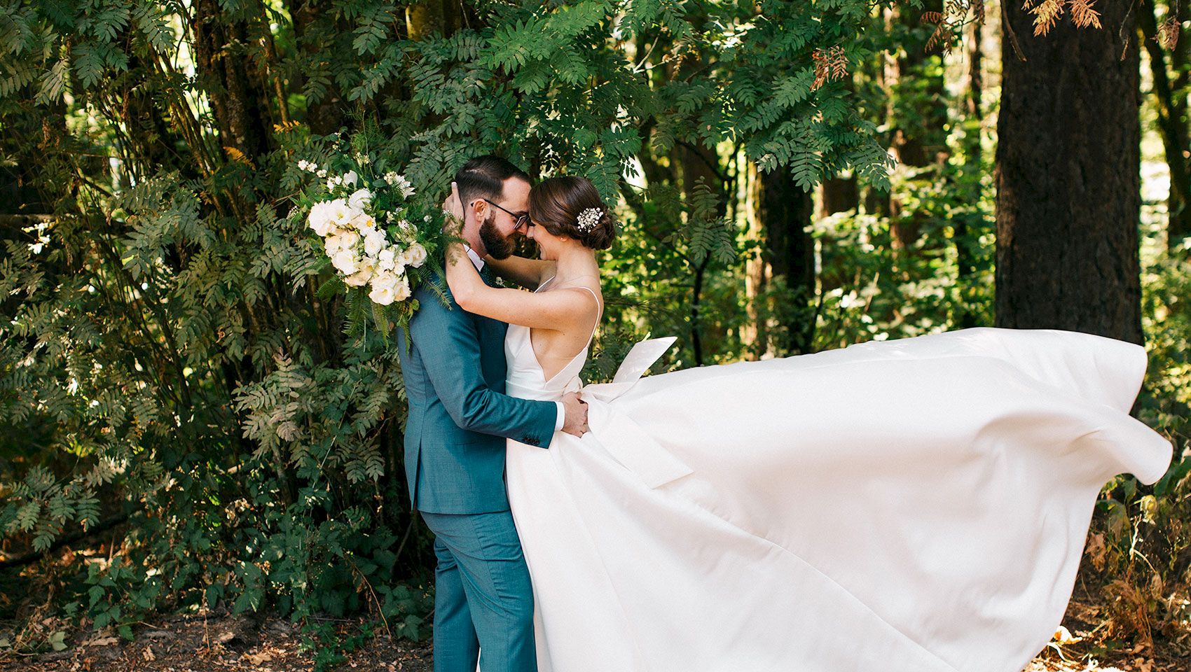 In a lush forest, a bride and groom in wedding attire share a kiss while holding white flowers.