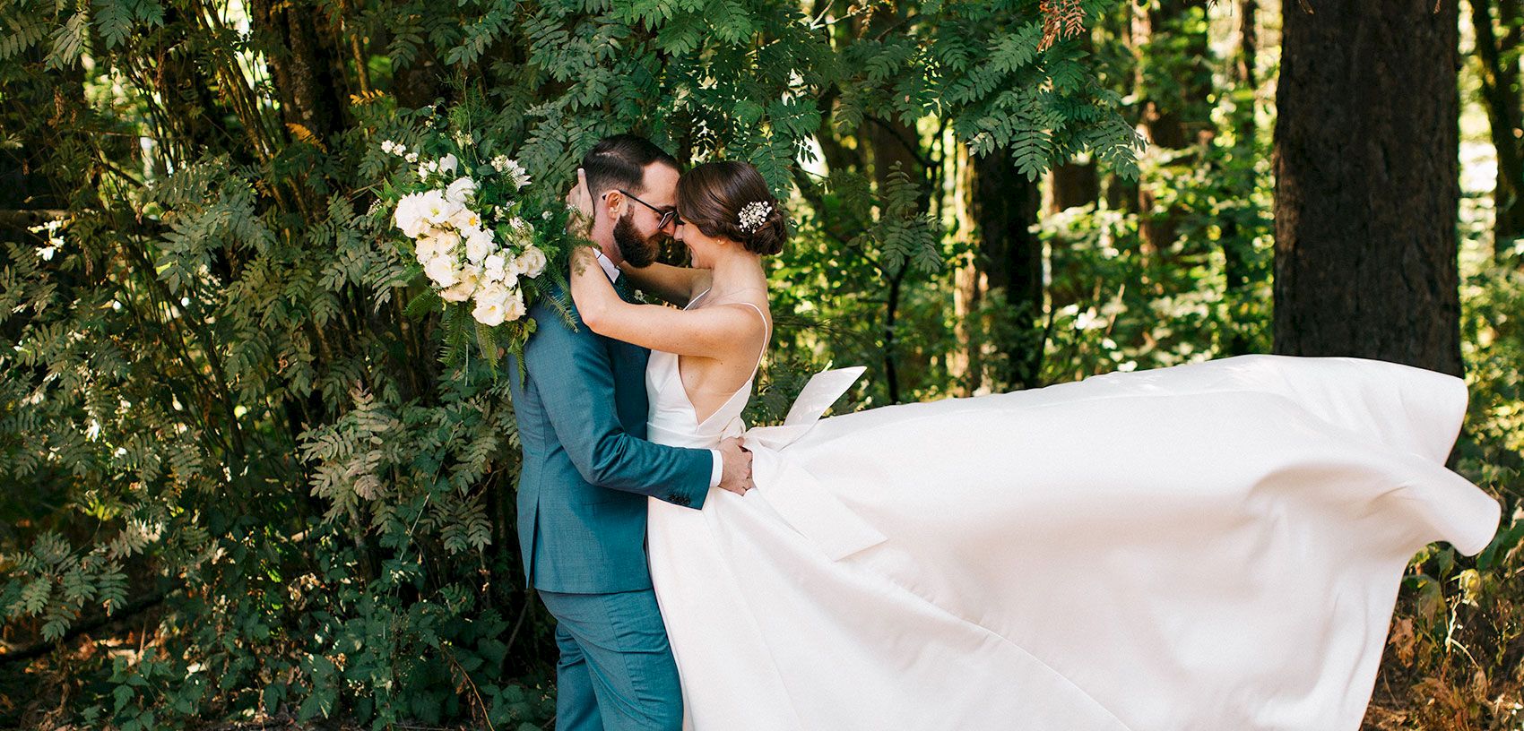 In a lush forest, a bride and groom in wedding attire share a kiss while holding white flowers.