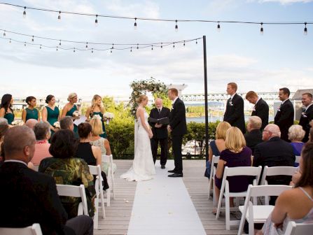 An outdoor wedding ceremony on a wooden deck, with the bride and groom exchanging vows under string lights, while guests watch.