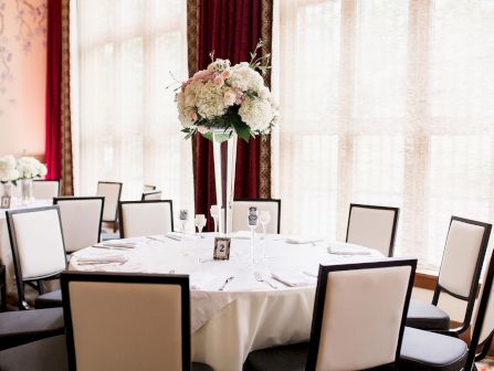 An elegant banquet table set with white place settings and a pink-and-white flower centerpiece against red curtains.