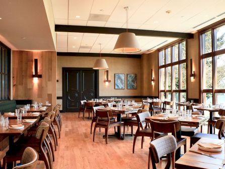 A modern restaurant dining room with wooden tables, upholstered chairs, pendant lights, and large windows letting in daylight.