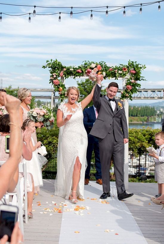 An outdoor wedding ceremony: bride and groom stand beneath a floral arch on a deck while guests cheer and throw confetti.