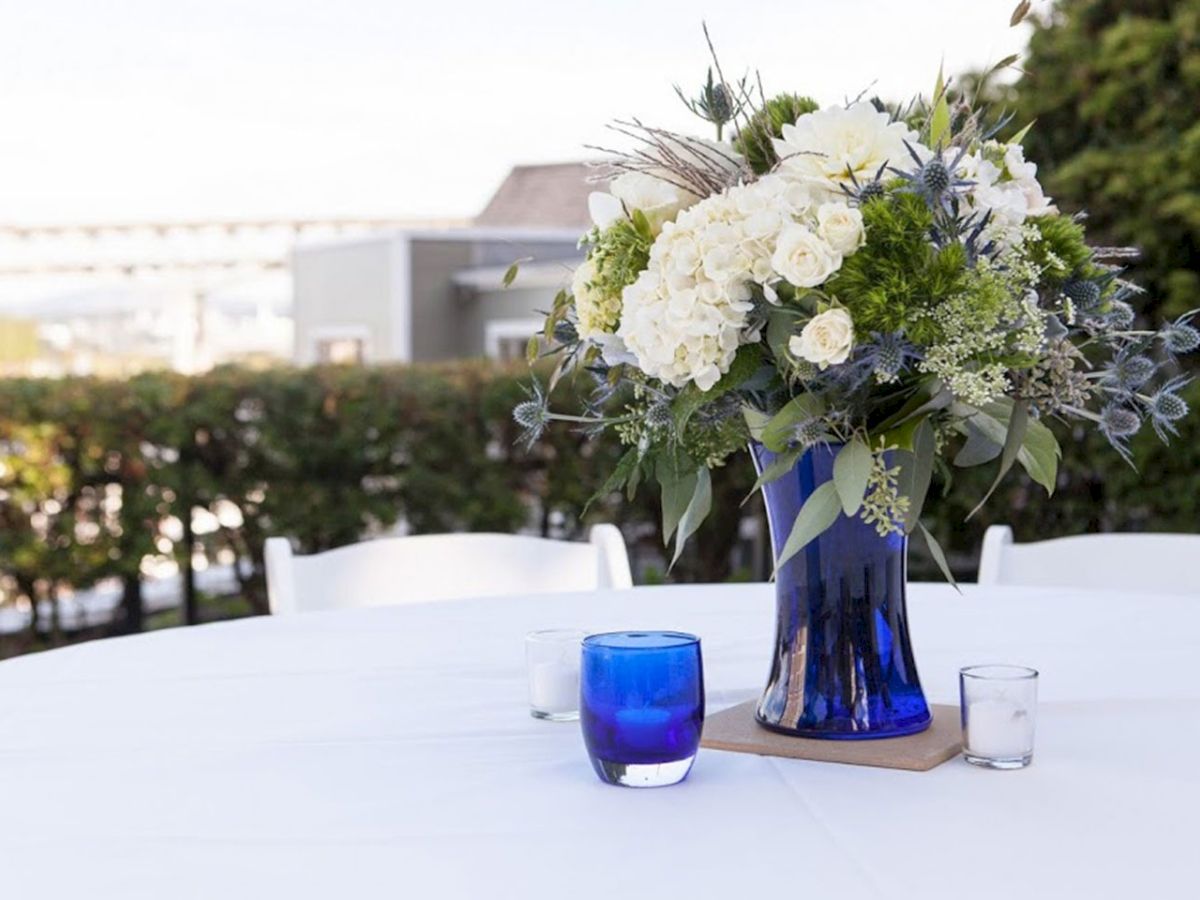 A white-and-blue floral centerpiece in a vase sits on an outdoor wedding-style table with glasses in front.