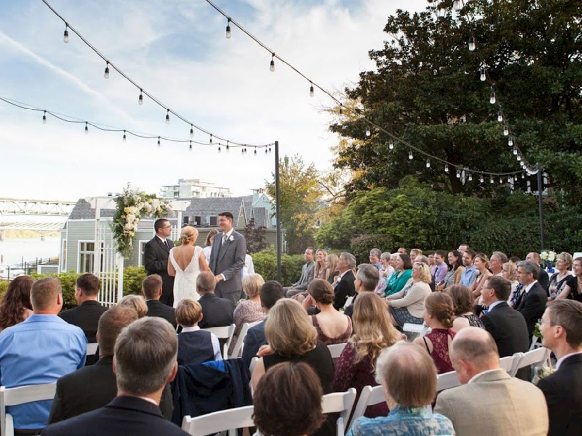 An outdoor wedding ceremony on a terrace with a bride and groom at the front, guests seated in rows, and string lights overhead.