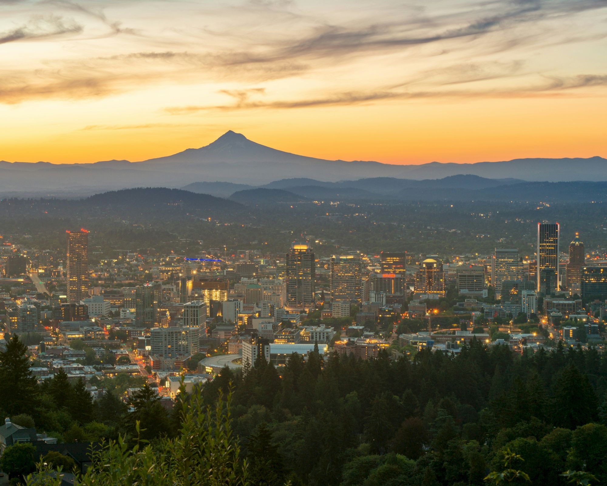 City skyline at sunset with a large mountain in the background and glowing lights across the valley.