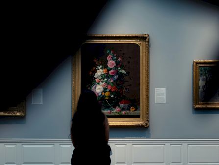 A museum visitor sits at a bench studying a framed bouquet painting on a gallery wall under spotlights.