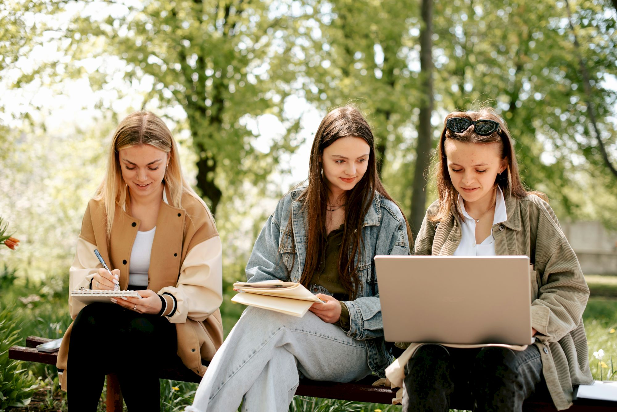 Three young women study outdoors on a bench, one writing in a notebook while another reviews papers and one uses a laptop.