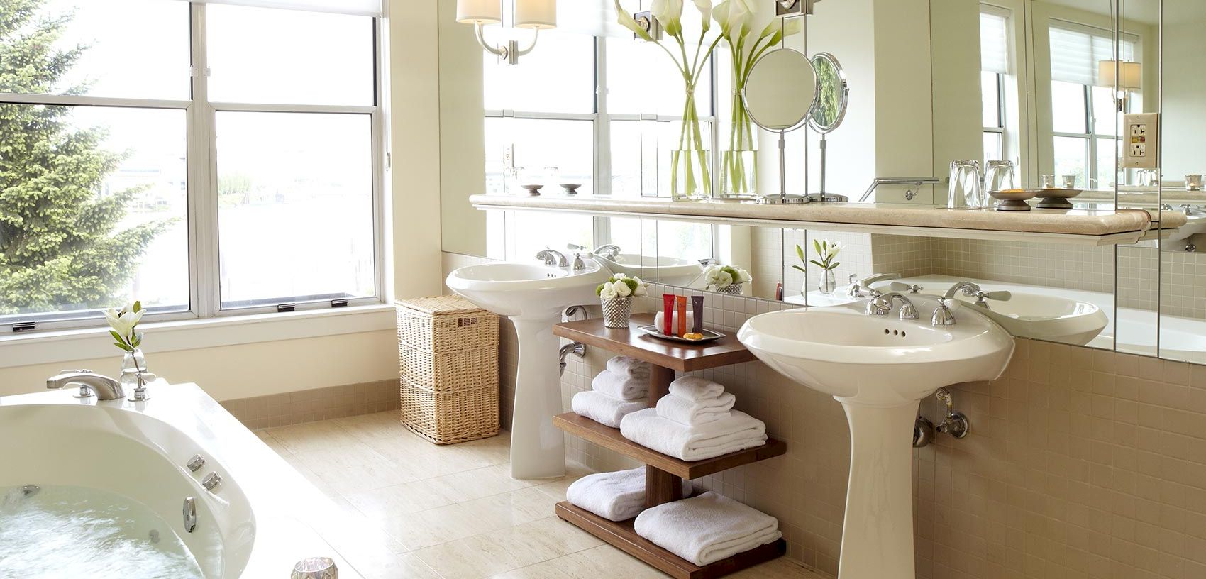 A bright, elegant bathroom with twin sinks, a bathtub, fresh flowers, and neatly stacked white towels by large windows.