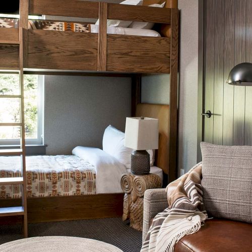 A stylish hotel room with stacked bunk beds, a patterned lamp on a nightstand, and a leather sofa near the window.