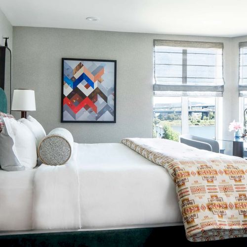 A bright modern bedroom with a made-up bed, throw pillows, framed art, and two large windows with blinds.