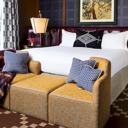 A stylish hotel bedroom with a made bed, patterned pillows, two wall lamps, and a cozy upholstered bench by windows.