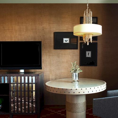 A modern living room with a flat-screen TV, sideboard, lamps, and a round table with a vase of flowers.